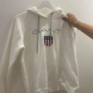 Vit hoodie från GANT - Vit hoodie från GANT med ett tryck av märkesnamnet och en sköld med stjärnor och ränder på bröstet. Tröjan har en justerbar huva med dragsko och långa ärmar.den har ett märke på sig