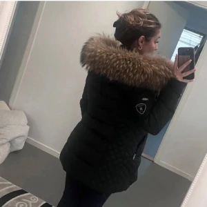 Svart dunjacka med päls från Rockandblue - Snygg svart dunjacka från Rockandblue med avtagbar päls på huvan. Jackan har quiltade detaljer och flera praktiska fickor med dragkedjor. Perfekt för kalla dagar med sin långa design och varma material.