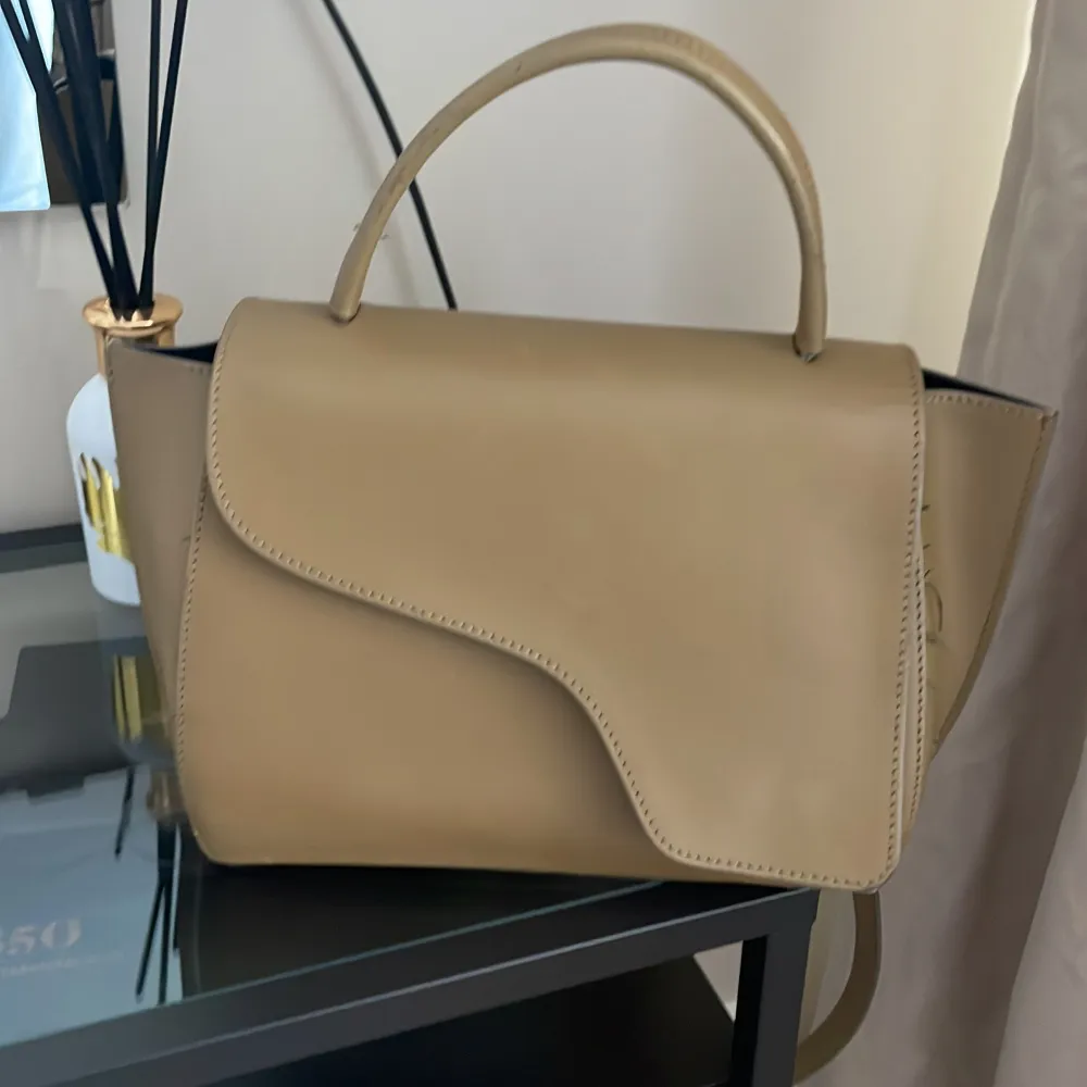 Snygg beige handväska i skinn med en unik asymmetrisk flikdesign. Väskan har ett handtag och en avtagbar axelrem för flexibilitet. Perfekt för att ge din outfit en stilren touch.. Laukut & Käsilaukut.
