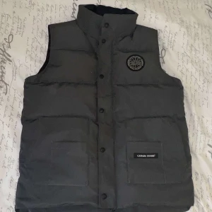 Svart dunväst från Canada Goose - Säljer en svart dunväst från Canada Goose med broderad logotyp på bröstet. Västen har knappar och är perfekt för kyligare väder. Den är tillverkad i polyester och nylon med fyllning av dun och fjädrar.