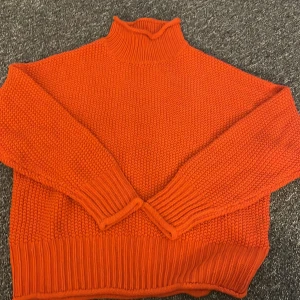 Orange stickad polotröja - Säljer en snygg orange stickad polotröja med hög krage. Tröjan har långa ärmar och ribbade muddar vid ärmslut och nederkant. Perfekt för kyliga dagar!
