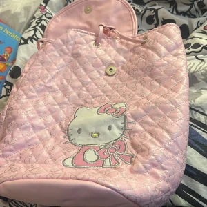 Rosa quiltad ryggsäck med Hello Kitty-motiv - Söt rosa ryggsäck med quiltat mönster och ett stort Hello Kitty-tryck på framsidan. Väskan har en dragkedja och justerbara axelremmar för bekväm passform. Perfekt för alla Hello Kitty-fans!