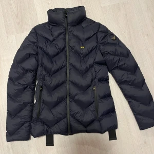 Oanvänd Svart dunjacka från Fendi - Snygg svart dunjacka från Fendi med dragkedja och hög krage. Jackan har bara hängt i garderoben sen ny. Jackan har ett quiltat mönster och justerbara remmar på baksidan. Perfekt för kyliga dagar.