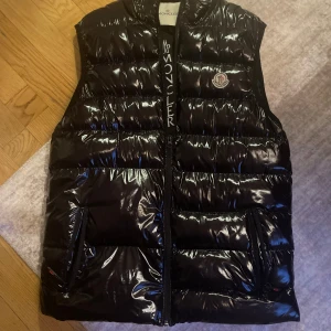 Svart dunväst från Moncler - Säljer en snygg svart dunväst från Moncler med glansig yta och dragkedja framtill. Västen har två praktiska fickor och Moncler-logga på bröstet. Perfekt för kyliga dagar!