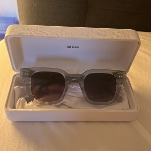 Chimi glasögon 04 grey  - Snygga grå solglasögon från Chimi med en fyrkantig form. De har en transparent grå båge och mörka linser, perfekt för en stilren look. Kommer i ett elegant vitt fodral med Chimi-logga.