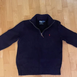 Mörkblå stickad tröja från Ralph Lauren - Säljer en snygg mörkblå stickad tröja från Ralph Lauren med dragkedja och ribbad krage. Tröjan har en liten röd logga på bröstet och är perfekt för kyligare dagar.