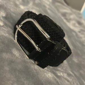 Svart flätat bälte med silverfärgat spänne - Snyggt svart flätat bälte med ett stilrent silverfärgat spänne. Perfekt för att ge en extra touch till din outfit. Bältet har en klassisk design som passar till många olika stilar.