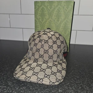 Beige keps med monogrammönster - Snygg beige keps från Gucci med det ikoniska monogrammönstret i svart. Kepsen har en justerbar rem baktill för perfekt passform och är tillverkad i ett slitstarkt material. Perfekt för att ge din outfit en lyxig touch. Snabb affär = billigare