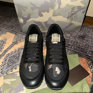 Valentino Garavani sneakers i svart och kamouflage - Snygga Valentino Garavani sneakers i svart med kamouflagemönster. Skorna har en klassisk snörning och en robust sula. Perfekta för en stilren look med en touch av edge.