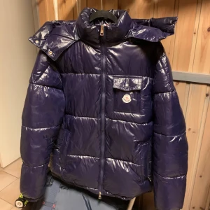 Mörklila pufferjacka från Moncler - Snygg mörkblå pufferjacka från Moncler med glansig finish. Jackan har en praktisk dragkedja och en ikonisk logotyp på bröstet. Den är långärmad och perfekt för kyligare väder. Storlek XL. - OM ANNONSEN ÄR KVAR FINNS JACKAN KVAR. 