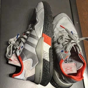 Grå och röda sneakers från Adidas - Snygga grå sneakers med röda och vita detaljer från Adidas. Skorna har en modern design med snörning och en bekväm sula. Perfekta för en sportig look. Modellen heter Nite Jogger. 