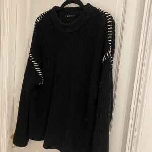 Svart stickad tröja från Gina Tricot - Snygg svart stickad tröja från Gina Tricot med unika vita detaljer längs ärmarna. Perfekt för kyliga dagar med sin mysiga och stilrena design. Tröjan har långa ärmar och en rund halsringning.