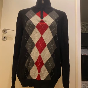 Svart tröja med argylemönster från Dressmann - Snygg svart tröja från Dressmann med klassiskt argylemönster i rött, grått och vitt. Tröjan har en dragkedja vid halsen och är tillverkad i 100% bomull. Perfekt för en stilren look.  Längd: 64 cm Armhåla till armhåla: 47 cm Trådsläpp, se bild!  Inga bilder när den sitter på mig! Katter finns i hemmet