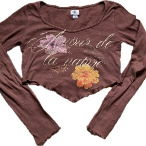 Brun croppad topp med blommotiv - Säljer en oanvänd  brun croppad topp från Uban Outfiters men köpt genom Plick, med texten 'Amour de la nature' och blommotiv i rosa och gult. Toppen har långa ärmar och en vågig nederkant. Perfekt för en avslappnad stil. 
