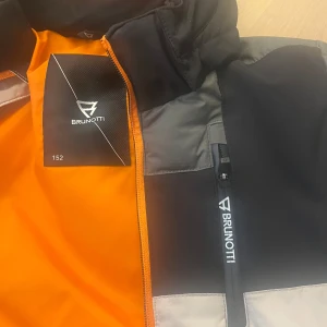 Beige/ svart skidjacka från Brunotti, storlek 152 - Säljer en snygg skidjacka från Brunotti i beige/grönt och svart med orange detaljer. Jackan har en dragkedja framtill och liftkortshållare både fram och på ärm. 2024 års modell Sanclairy Boys Puffer Snow Jacket från Brunotti 