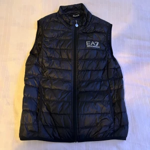 Svart dunväst från EA7 - Snygg svart dunväst från EA7 med dragkedja framtill. Perfekt för varma dagar. Västen har en quiltad design och en stilren logga på bröstet. Det står M på jackan men jag skulle säga att storleken ligger mellan S och M. Pris kan diskuteras vid snabb affär😀