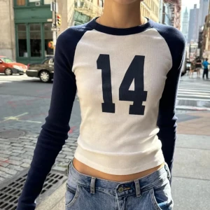 Vit och marinblå långärmad topp med nummertryck - Snygg och trendig vit topp med marinblå ärmar och stort nummertryck '14' på framsidan. Perfekt för en avslappnad stil. Den är långärmad och har en tight passform.
