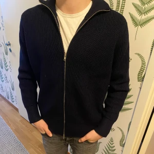 Maison Margiela full zip - Säljer en stilren mörkblå ribbad tröja från Maison Margiela i ny skick i storlek M Tröjan har en dragkedja framtill och långa ärmar.Den är tillverkad i ett mjukt och bekvämt material som ger en skön passform. 