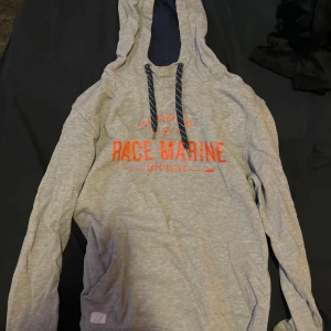 Grå hoodie från Race Marine (SWISH ENDAST!) - Säljer en grå hoodie från Race Marine med röd text på bröstet. Tröjan har en justerbar huva med snören och långa ärmar. Perfekt för en avslappnad stil.