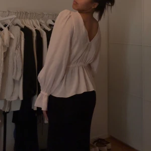 Vit blus med volangdetaljer - Elegant vit blus med långa ärmar och volangdetaljer vid midjan. Blusen har en v-ringad rygg och elastisk midja för en smickrande passform. Perfekt för en stilren look.