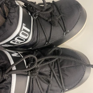 Svarta Moon Boots storlek 33-35 - Snygga och varma svarta Moon Boots med snörning och vit logga. Perfekta för kalla vinterdagar med sin unika design och komfort. Knappast använda😊