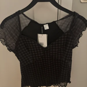 Svart och vit rutig topp från H&M Divided - Snygg svart och vit rutig topp från H&M Divided med korta ärmar och volangdetaljer. Toppen har en transparent meshdesign och en croppad passform. Perfekt för en trendig look!