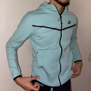 Ljusblå Nike tech - Snygg ljusblå huvtröja från Nike med dragkedja och svart logotyp på bröstet. Tröjan har en huva och långa ärmar, perfekt för en sportig look. Tillverkad i mjukt material för extra komfort. Ny skick, användes några gånger, storlek L men är liten i storlek, kan passa S eller M