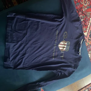 Mörkblå långärmad tröja från GANT - Säljer en stilren mörkblå långärmad tröja från GANT med tryck på framsidan. Perfekt för en avslappnad och snygg look. Tröjan har en rund halsringning och är tillverkad i ett mjukt material.