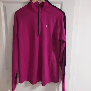 Rosa tränings tröja - Säljer en rosa Dri-FIT tröja från Nike, helt ny! Detalj med halv dragkedja och långärmad design. Perfekt för löpning eller träning med sin fukttransporterande teknologi. Svarta detaljer längs ärmarna och en liten Nike-logga på bröstet.