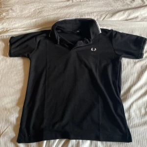 Svart pikétröja från Fred Perry - Snygg svart pikétröja från Fred Perry med klassisk krage och vit broderad logga på bröstet. Perfekt för en stilren och avslappnad look. Kragen har en vit kant som ger en extra touch.