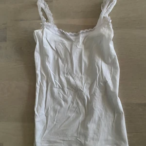 Vit linne med spetsdetaljer - Säljer ett stilrent vitt linne med spetsdetaljer vid axelbanden och halsringningen. Perfekt för varma sommardagar eller som ett basplagg i garderoben. Aldrig använd 
