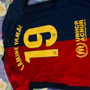 FC Barcelona fotbollströja - Snygg FC Barcelona tröja från Nike med korta ärmar. Tröjan är i rött och blått med gula detaljer, inklusive klubbens emblem och sponsorloggor. Perfekt för fans som vill visa sitt stöd till deb unga killen Lamine Yamal.