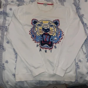 Vit sweatshirt från Kenzo med tigerbroderi - Säljer en vit Kenzo sweatshirt med ett stort, färgglatt tigerbroderi på framsidan. Tröjan har långa ärmar och en rund halsringning. Perfekt för en stilren och trendig look.