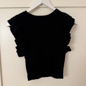 Svart ärmlös topp med volangdetaljer - En gullig svart croppad topp med volanger runt armarna/axlarna från Even&Odd. Användts flitigt men är tyvärr för liten. Den är av stretchigt material därav de vita prickarna/trådarna som syns uppe vid volangerna som blivit efter användning och tvättning. Utöver det är toppen i fint skick. Sitter mer som XS än S🖤Nypris: 249kr