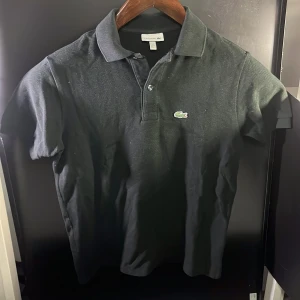 Svart pikétröja från Lacoste - Säljer en klassisk svart pikétröja från Lacoste med korta ärmar och den ikoniska krokodilloggan på bröstet. Perfekt för en stilren och avslappnad look. Hör av er vid minsta fundering🙌