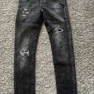 Jack & Jones Svarta jeans med slitningar  - Snygga svarta jeans från Jack and Jones med coola slitningar på framsidan. De har en tight passform och klassisk femficksdesign. Perfekta för en avslappnad stil med en edgy touch.