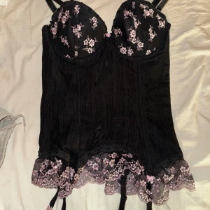 Svart korsett med rosa blommor - Supergullig  svart korsett med rosa blommönster och spetsdetaljer. Den har justerbara axelband och en gullig spetskant längst ner. Osäker på storlek men säger S-M, aldrig använd🩷avtagbara snören längst ner 