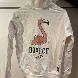 Vit hoodie från Dope - Säljer en vit hoodie från Dope med en cool flamingo-grafik på baksidan och texten 'VACAY'. Den har en känguruficka och justerbar huva med dragsko. Perfekt för en avslappnad stil! Något liten i storleken. 