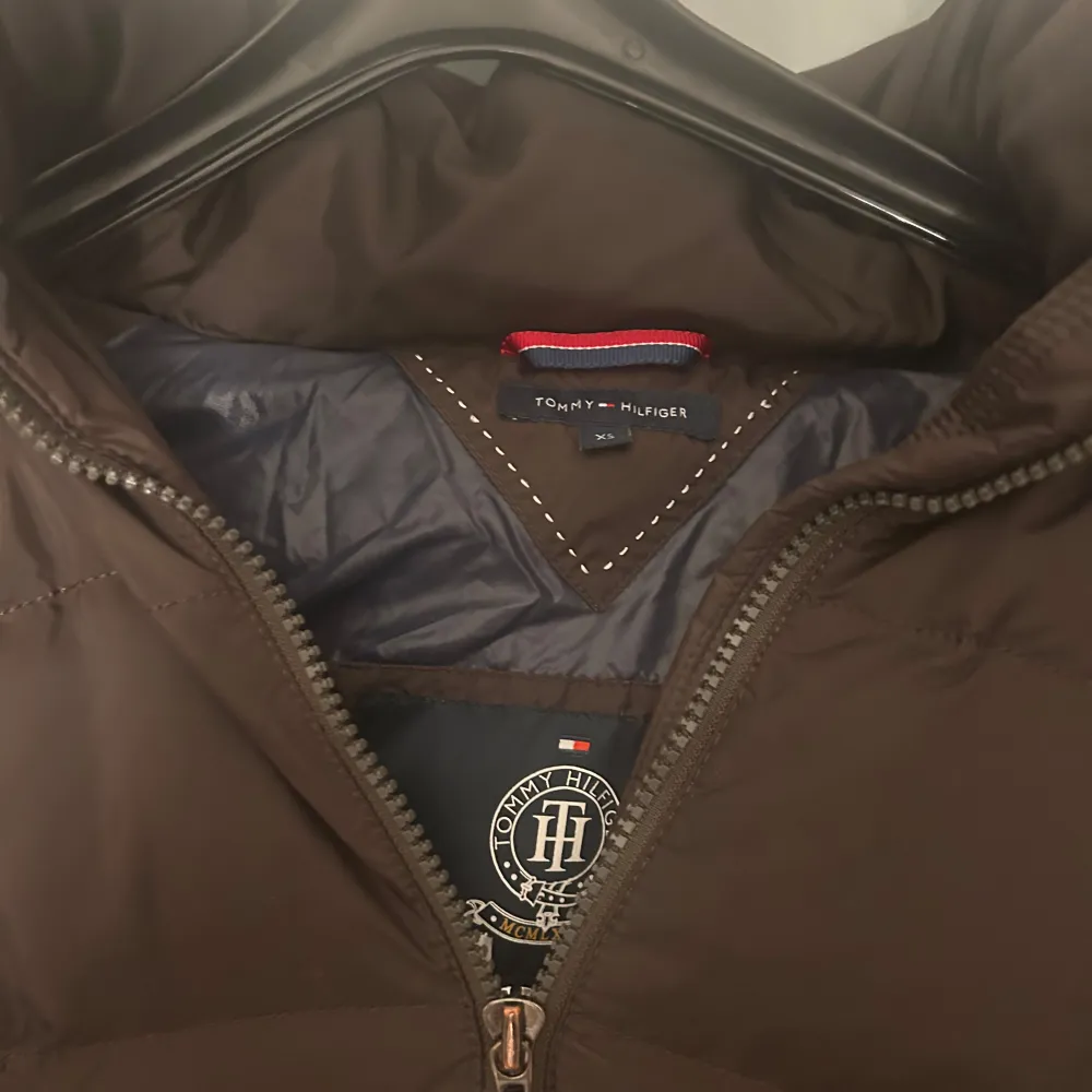 Säljer en brun dunjacka från Tommy Hilfiger med pälskantad huva och dragkedja framtill. Jackan har en quiltad design och praktiska fickor med knappar. Perfekt för kyliga dagar.. Takit.
