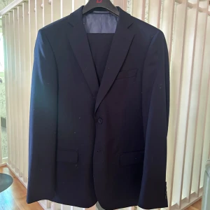 Marinblå kostym - Elegant marinblå kostym med kavaj och byxor. Kavajen har en klassisk design med knappar framtill och fickor. Perfekt för formella tillfällen. Strl 50(M/L) i kavajen, strl 33/32 i byxorna. Använd 2 gånger, ser ut precis som ny!