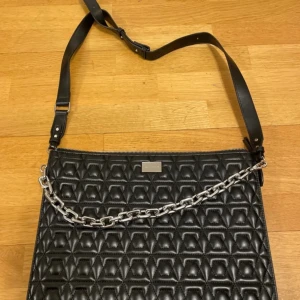 Svart quiltad axelväska med kedjedetalj - Snygg svart axelväska/ datorväska i quiltat mönster med en cool kedjedetalj som är avtagbar. Väskan har en justerbar axelrem och stängs med dragkedja. Använts två gånger, är i princip helt i helt ny skick. Mått: 27 x 37cm.