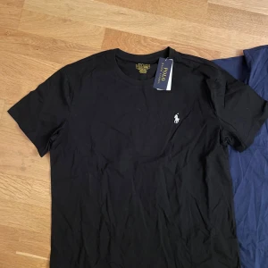 Svart t-shirt från Polo Ralph Lauren - Snygg svart t-shirt från Polo Ralph Lauren med klassisk passform. Den har en liten broderad logga på bröstet och är kortärmad. Perfekt för en stilren look.