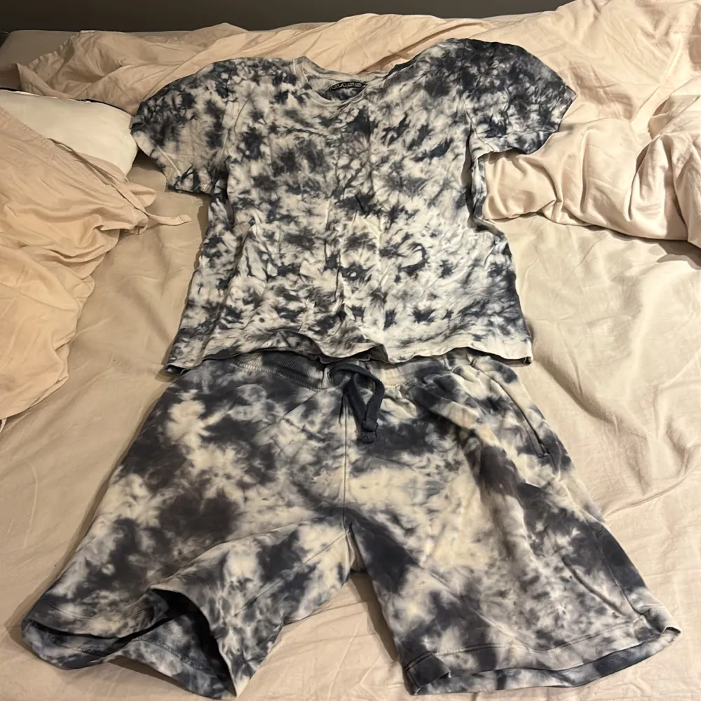Säljer en cool svartvit batikmönstrad t-shirt med matchande shorts. T-shirten är kortärmad och setet är perfekt för en avslappnad stil. Materialet ser mjukt och bekvämt ut, idealiskt för varma dagar. Är samma storlek på T-shirten och shortsen.. T-paidat.