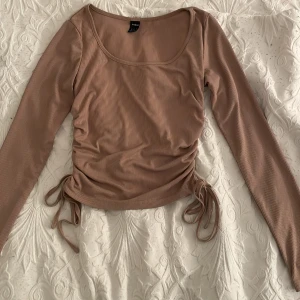 Beige ribbad topp med knyt - Säljer en beige ribbad topp med långa ärmar och knytning i sidorna. Perfekt för en stilren look. Toppen har en rund halsringning och är i ett mjukt material.