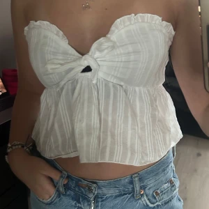 Vit randig topp från SHEIN - Säljer en söt vit randig topp från SHEIN med knytning framtill. Toppen är axelbandslös och har en volangdetalj vid kanten. Perfekt för varma sommardagar!