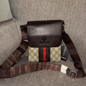 Gucci axelväska med monogram - Snygg axelväska från Gucci med klassiskt monogrammönster. Väskan har en brun läderdetalj på locket och en justerbar axelrem med Gucci-logga. Den har en dragkedja på baksidan och ett huvudfack med dragkedja. Perfekt för att bära dina nödvändigheter med stil.