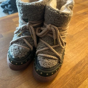 Gröna och beiga Inukkis med snörning och nitar - Säljer ett par varma och stiliga boots från Inuikii i Grön och brun färg. Skorna har en mysig pälsliknande yta och är dekorerade med snörning och nitar. Perfekta för kyliga dagar.