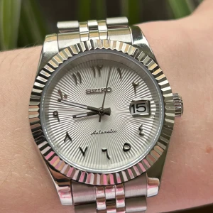 Seiko Mod Arabic-Dial ”Datejust” - Snygg automatisk seikomod med silverfärgad boett och armband, boett är 40mm bred. Urtavlan har ett elegant mönster och tydliga arabiska siffror. Datumvisning vid klockan tre och transparent bakboett som visar urverket. Inuti sitter ett nh35 urverk från seiko. Grön lume som lyser i mörker. Mejsel och klockkudde medföljer.    Skicka Dm vid frågor eller prisförslag 😁📩