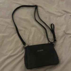 Svart axelväska från Guess - Snygg svart axelväska från Guess med justerbar rem och flera fack med dragkedja. Perfekt för att hålla ordning på dina saker. Väskan har en stilren design med Guess-loggan i silver på framsidan.