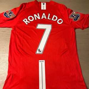 Röd Manchester United fotbollströja - Säljer en röd Manchester United fotbollströja med 'Ronaldo' och nummer 7 på ryggen. Tröjan har korta ärmar och Premier League-märken på båda ärmarna. Framsidan pryds av AIG-loggan och klubbmärket. Perfekt för fans av laget och spelaren!